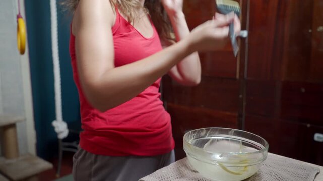 A determined woman raises her arm to dye her hair next to a mixture bowl in her cozy room. She feels hopeful and focused on her self-care routine.