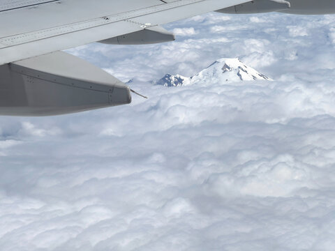 airplane wing and aerial view of heavy cloud cover and mostly snow-covered peak of Mt. Baker in Mt. Baker-Snoqualmie National Forest, National Park, viewed from a jet in July 2026