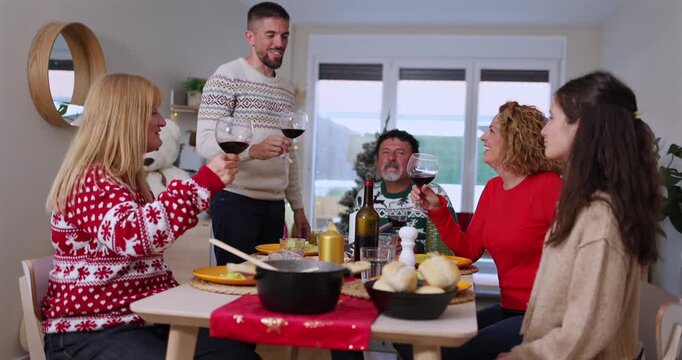 Cheerful multi generational family wearing festive sweaters making a toast with glasses of red wine during a christmas holiday dinner, smiling and celebrating together at home