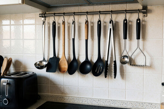 Set of metal, wood kitchen tools hang on wall rail above modern stove. Black spatula ladle spoon tongs masher accessory for daily cooking meal prep routine. Home interior design with ceramic tiles,