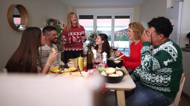 Happy multiethnic family enjoying christmas dinner together at home, a senior woman makes a toast with red wine while everyone listens and smiles in their festive sweaters at the dining table