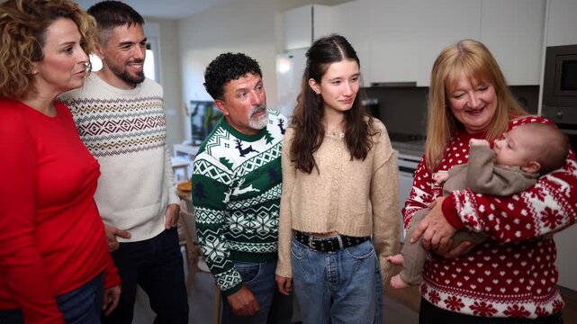 Multigenerational family gathering at home for christmas, joyfully laughing and admiring the new baby held by the smiling grandmother, all wearing festive holiday sweaters in a cozy atmosphere