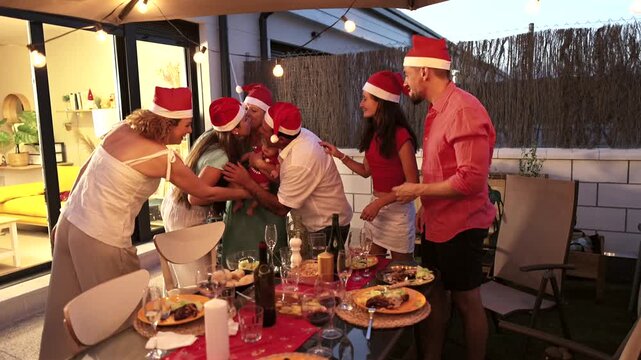 Joyful multi generational family wearing santa hats gathering and hugging during a christmas eve dinner celebration on the patio, enjoying a festive holiday meal together under warm string lights