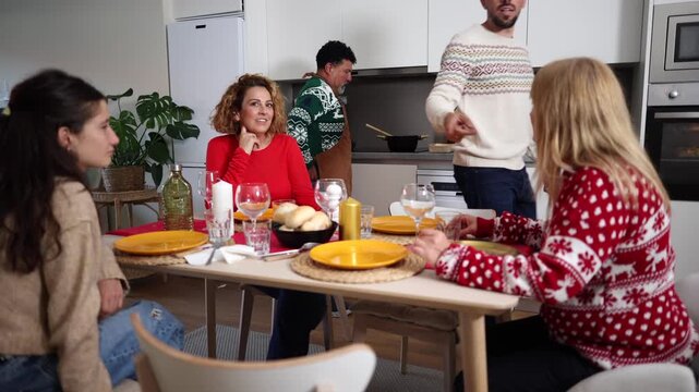 Multiracial group of friends wearing festive sweaters enjoying a conversation while sitting at a dining table during a christmas celebration at home, with a man cooking in the background