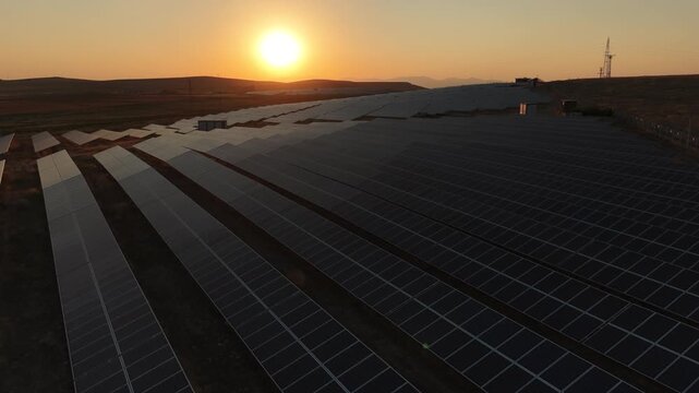 Cinematic aerial drone flight over a massive solar panel farm at sunset, renewable green energy concept.