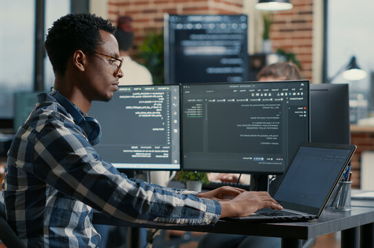 African American developer writes code on a laptop at a desk with multiple screens, working on user interface programming in a software agency.