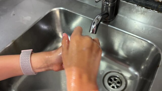 Close-up of child washing and scrubbing soapy hands under running water faucet in stainless steel kitchen sink