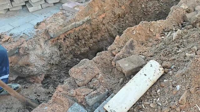 Individual using a shovel to dig a trench in sandy soil, revealing bricks and a white pipe, with a construction site visible in the background