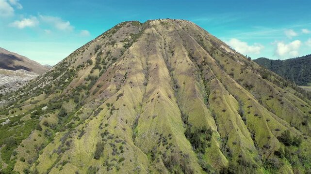 Aerial drone footage flying backwards away from the scenic Mount Batok volcano cone, showcasing the surrounding green landscape of East Java.