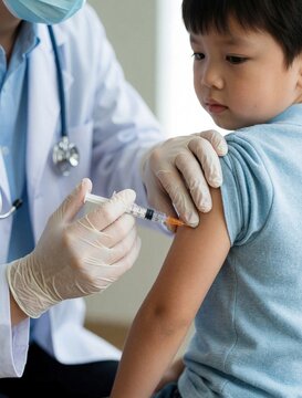 Pro doctor in white coat gives medical vaccine injection to little asian boy at clinic. Pediatrician uses sterile syringe for immunization procedure. Kid patient sits during health checkup session.