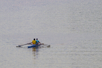 Two people row a small boat on calm water. They use oars to propel the vessel forward. The water is gentle with subtle ripples. The rowers wear bright tops for visibility. Their motion suggests steady © vefimov