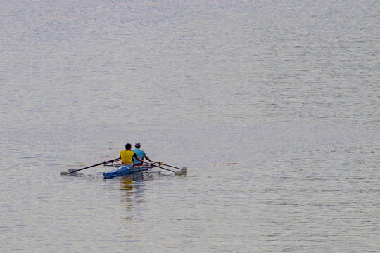 Two people row a small boat on calm water. They use oars to propel the vessel forward. The water is gentle with subtle ripples. The rowers wear bright tops for visibility. Their motion suggests steady