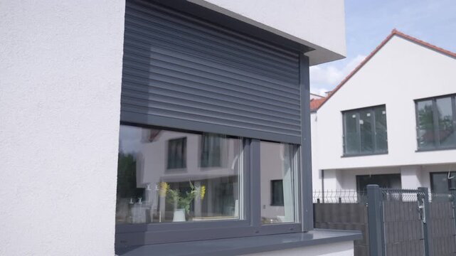 Dark gray roller shutter descends across modern residential window, gradually covering the glass. Concept of exterior shading and privacy