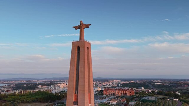 Aerial view of the monumental Cristo Rei statue rising above Almada, Portugal, illuminated by warm evening light beneath a blue sky with soft clouds.