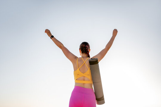 Woman holding yoga mat with arms raised at sunrise