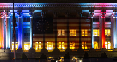 Obraz premium America flag covers federal building. America anniversary lights historic facade. Patriotic banner marks America freedom. Government columns frame America celebration.