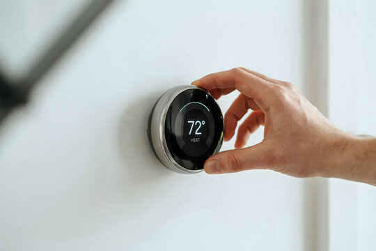 Close up of a hand rotating the outer ring of a round smart thermostat on a white wall to set the temperature to seventy-two degrees