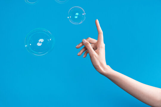 Caucasian woman hand reaching toward soap bubble on blue