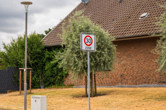 Speed limit 30 zone traffic sign installed on a metal pole in a quiet European suburban residential neighborhood next to a brick house and green tree.