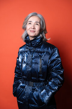 Senior Asian woman in glossy puffer coat on orange backdrop
