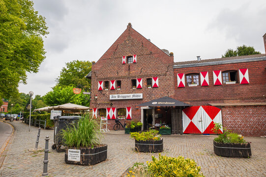 Traditional historic old brick watermill building facade with red and white shutters, cobblestone courtyard, outdoor restaurant terrace, and green trees on an overcast summer day