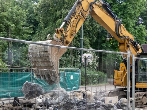A yellow excavator performing demolition, excavation work behind a temporary metal fence. Rubble, cleared debris are visible. A green mesh screen is attached to the fence to provide safety.