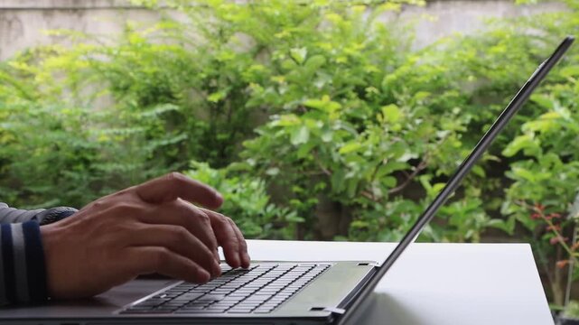 Man hand typing on computer keyboard. Businessman working with laptop on white table in garden at home office or workplace. Remote work, live chatting, online communication, Business technology.
