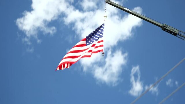 Large Red White and Blue American Flag Blowing in the Wind Hanging From Tall Crane Near Trees With Blue Sky