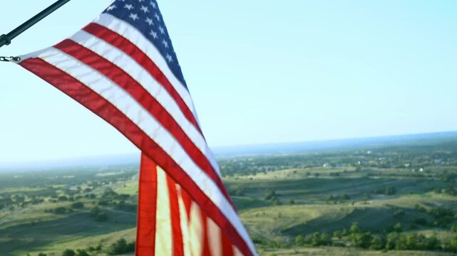 Large Red White and Blue American Flag Blowing in the Wind From Side of Tall Building Overlooking Rolling Green Hills With Blue Sunset Sky