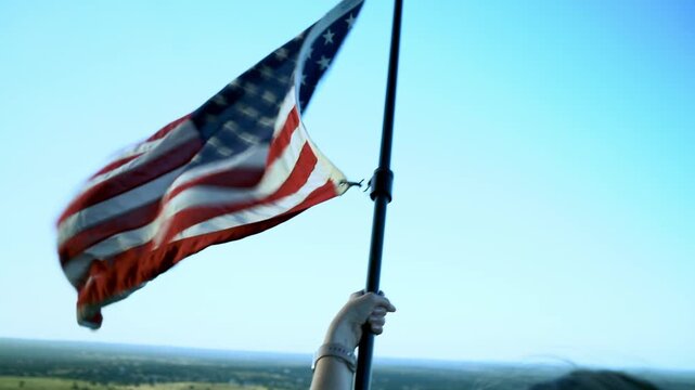 Large Red White and Blue American Flag Blowing in the Wind Hanging From Tall Crane Near Trees With Blue Sky