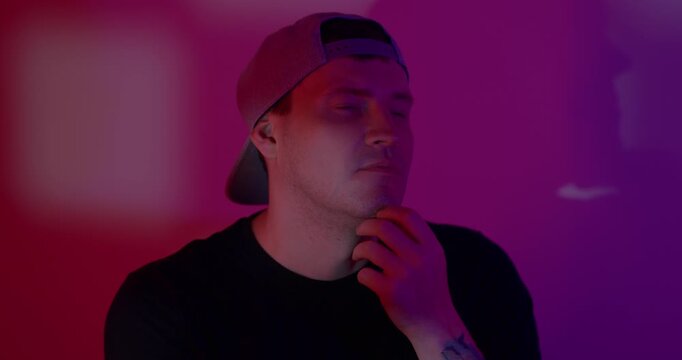 Man with cap thinking in neon light. Caucasian guy touching his chin with pensive expression. Portrait of thoughtful young male in dark studio with color glow.