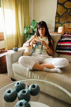 Young woman knitting on sofa in cozy sunlit living room