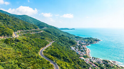 Obraz premium A scenic winding road cuts through green mountains beside a turquoise sea with a small coastal village under clear blue sky.