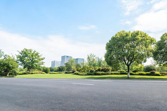 A wide asphalt road extends in the foreground, flanked by lush green trees and grass, with modern office buildings visible in the distance under a clear blue sky.