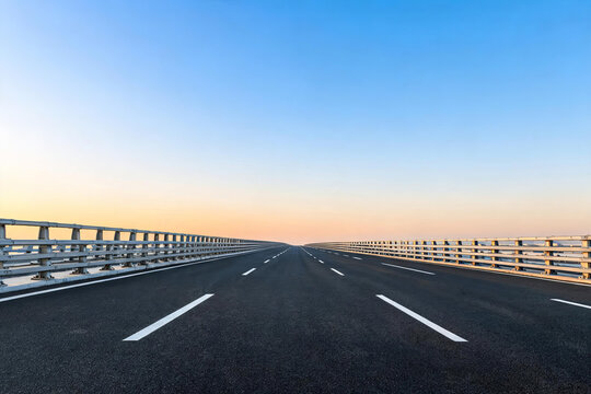 An empty asphalt bridge road stretches straight into the horizon, flanked by metal barriers under a clear gradient sky.