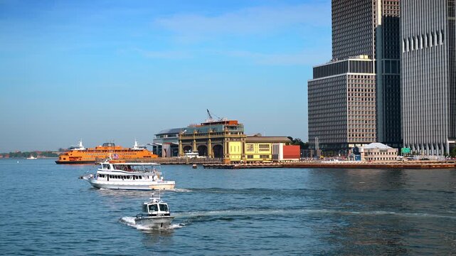 Tour boat and patrol craft near South Ferry. Two white vessels cross blue water before waterfront terminals and tall office towers.