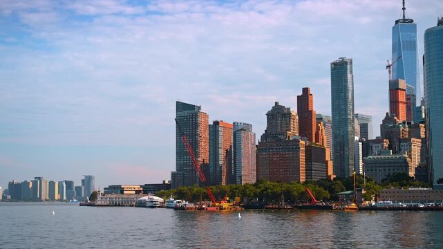 Lower Manhattan skyline above Hudson River waterfront. Cranes and varied towers catch warm sunlight beneath a partly cloudy sky.
