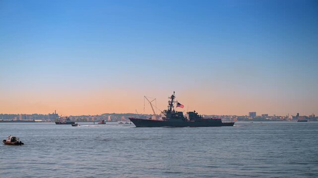 US Navy destroyer sailing through New York Harbor at dusk. Escort boats and a distant city silhouette frame the warship beneath an orange sunset sky.