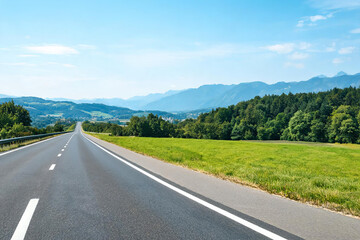 Fototapeta premium A straight empty asphalt road stretches into the distance, flanked by green grass and forests, with layered mountains under a clear blue sky.