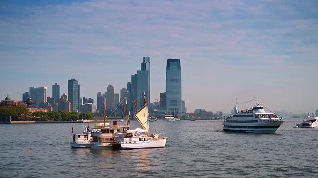 Classic yachts sailing near Jersey City skyline in New York Harbor. A passenger ferry crosses the water past modern towers under a soft afternoon light.