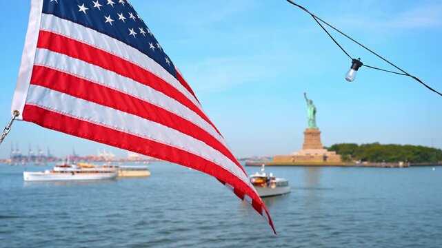 Frayed American flag hanging above New York Harbor near Statue of Liberty. Small boats drift near Liberty Island beneath a string of hanging lights and clear sky.