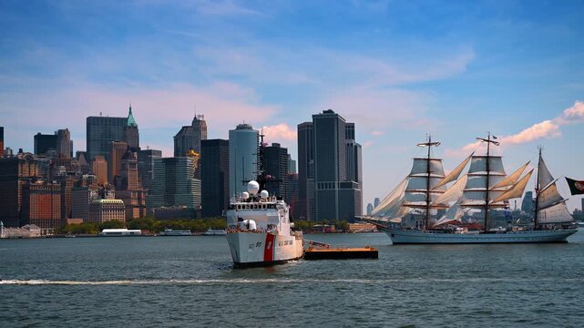 US Coast Guard cutter towing barge near tall ship in Manhattan. Full sails rise behind the vessels as historic towers frame the Lower Manhattan skyline.