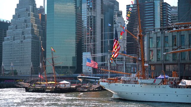 U.S. Coast Guard tall ship at South Street Seaport. American flags and signal pennants hang from orange masts before dense office towers.