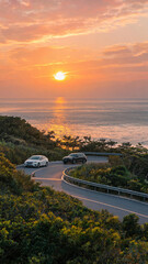 Obraz premium Two cars drive on a winding coastal road at sunset, with ocean and vibrant sky in the background.