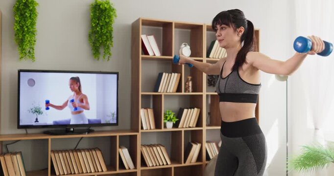 Woman home workout fitness online training exercise. Athletic adult uses dumbbells while following a tv class in a bright living room, building strength and toning arms. Clear concept remote fitness.