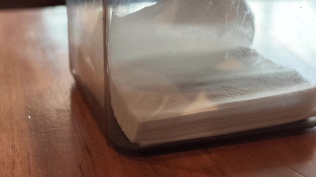 Macro slow motion shot focusing on the stack of folded white paper napkins stored inside a transparent plastic tissue dispenser box.
