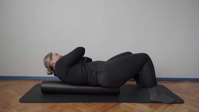 Plus size exerciser rocks side to side on a foam roller to massage the upper back in a quiet indoor room.