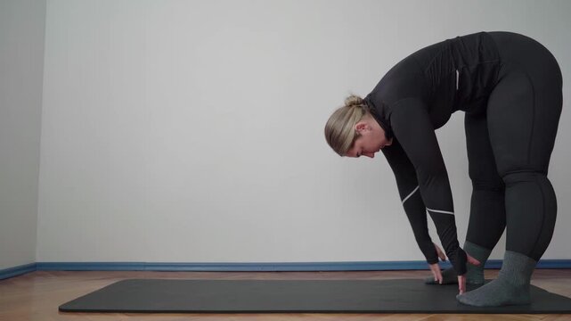 Adult exerciser walks hands into a forward fold on a mat, lengthening hamstrings and decompressing the spine in a plain indoor room.
