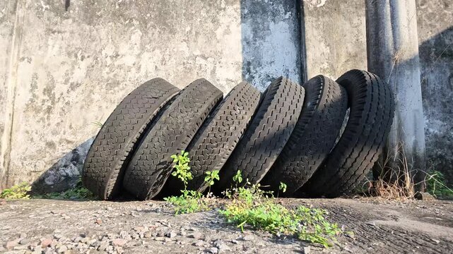 Wide slow motion shot of old black truck tires lined up leaning against a weathered concrete wall outdoor on sunny afternoon.
