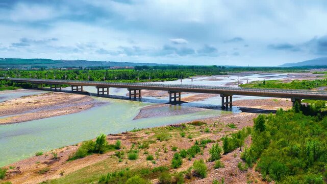 Aerial view of a long concrete bridge spanning a wide river valley surrounded by lush green foliage under a cloudy blue sky in Xinjiang.